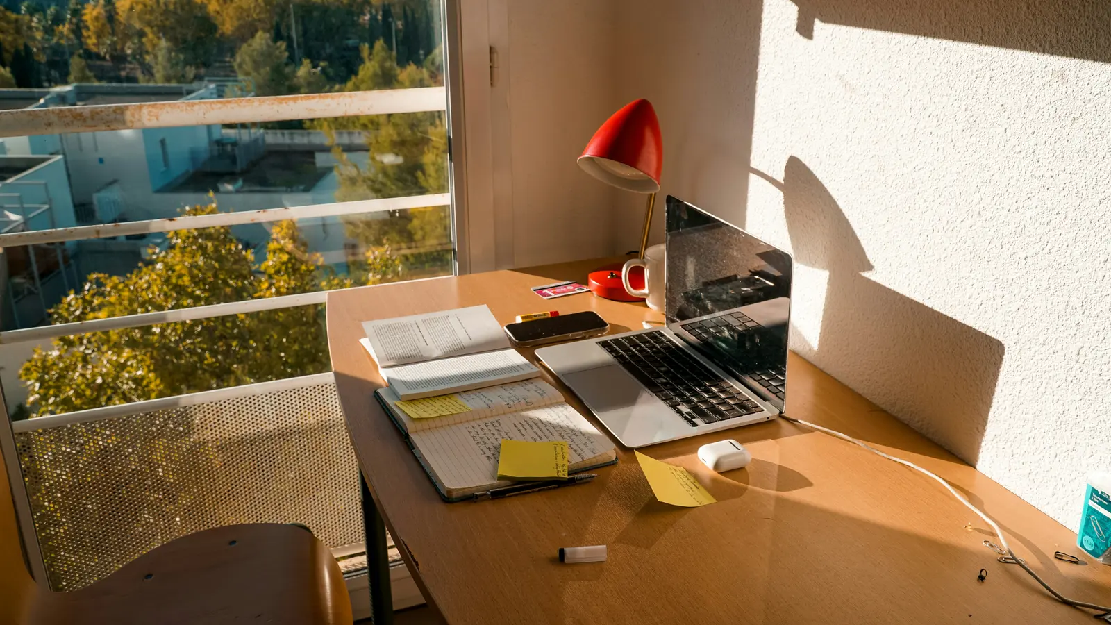 A laptop and notebook on a wooden desk beside a sunlit window.